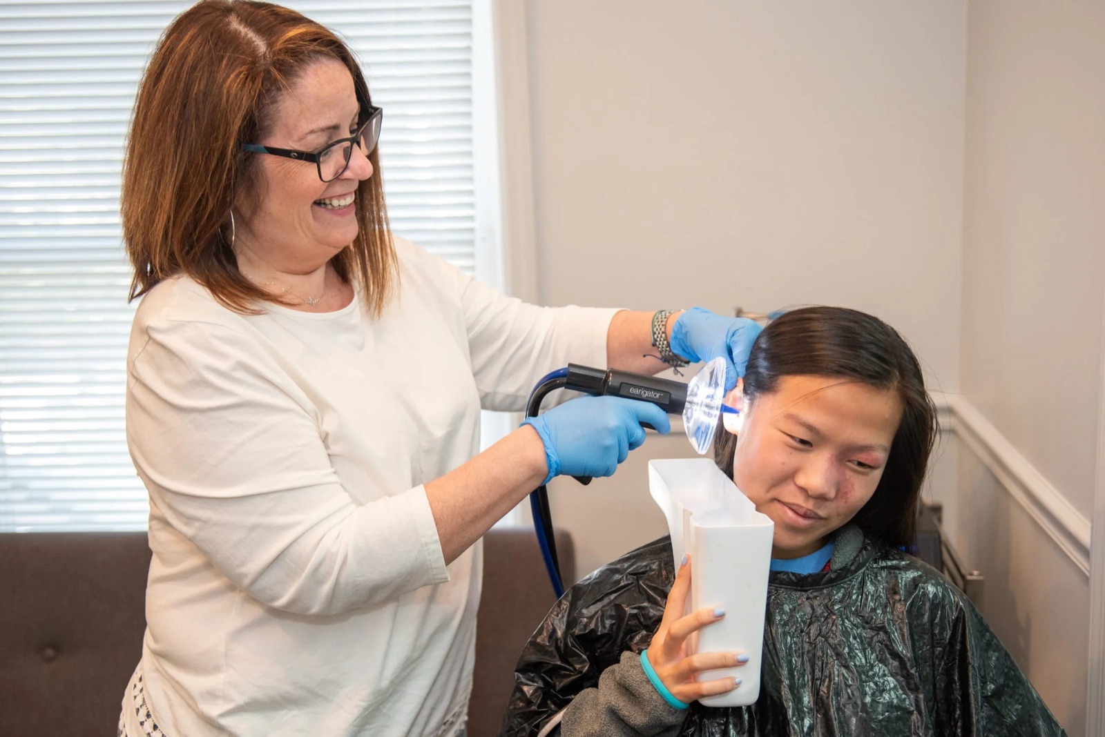 Dr. Kalady, in blue gloves, smiling as she rinses earwax from a young patient's ear while the patient holds a basin