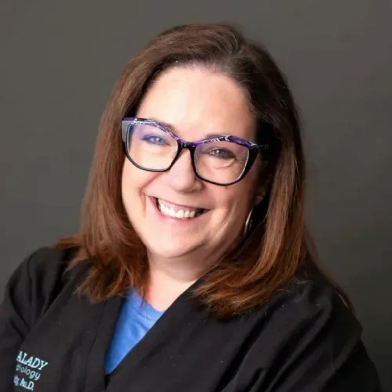 Dr. Meg Kalady, audiologist, smiling in purple-framed glasses and a black Kalady Audiology jacket over a blue top